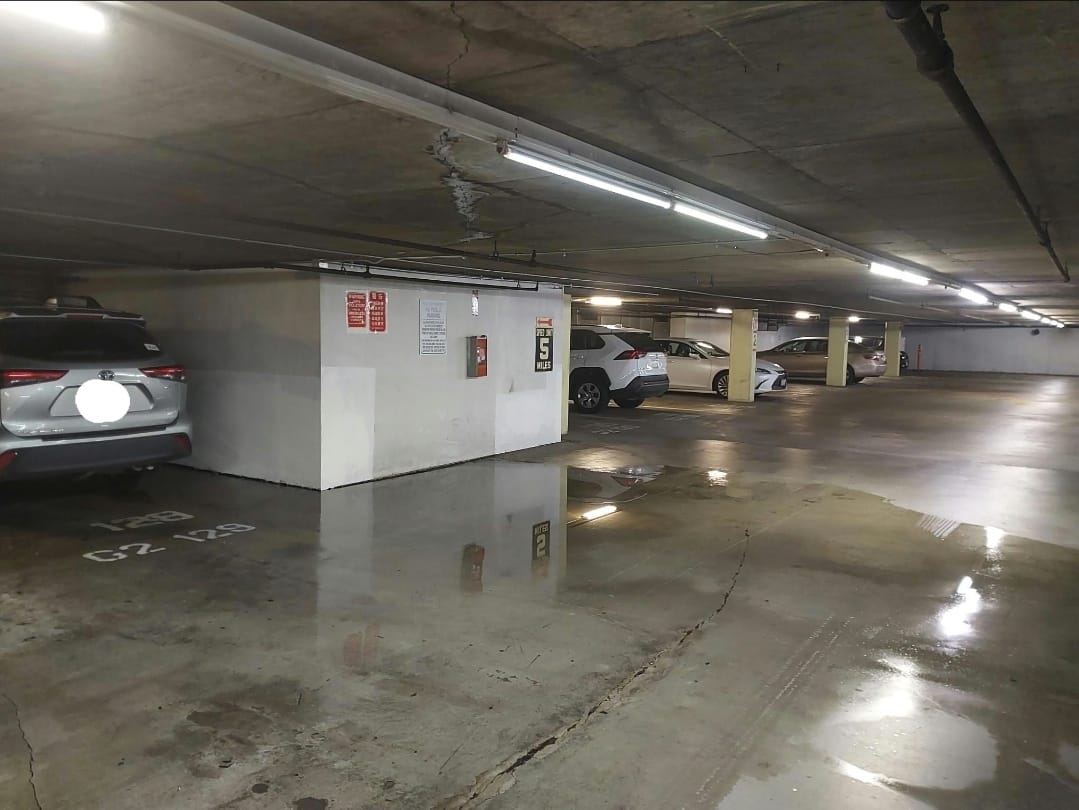Parking garage with standing water on the floor, ceiling cracks, and signs of concrete leakage issues.